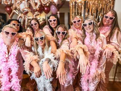 Group of women in pink and white robes with heart-shaped sunglasses, posing with arms stretched out showing their permanent bracelets at an Everlove private permanent jewelry party in Charleston, SC.